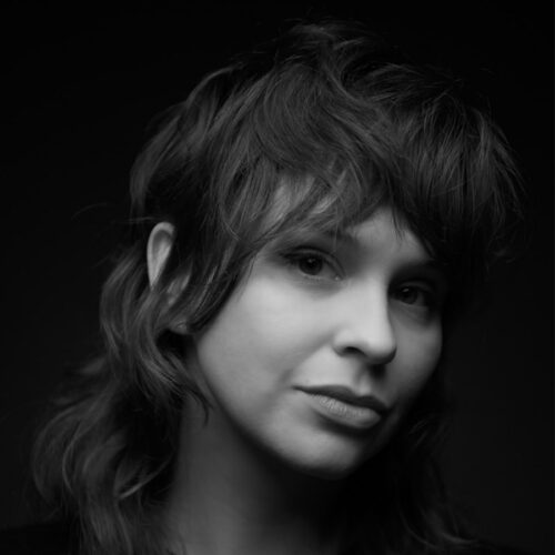Black and white portrait of a person with layered, shaggy hair, looking slightly to the side with a neutral expression against a dark background.