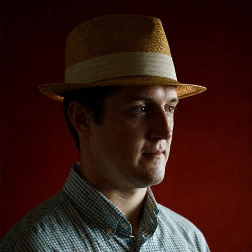 Man wearing a straw hat and a checkered shirt, facing sideways against a dark red background.