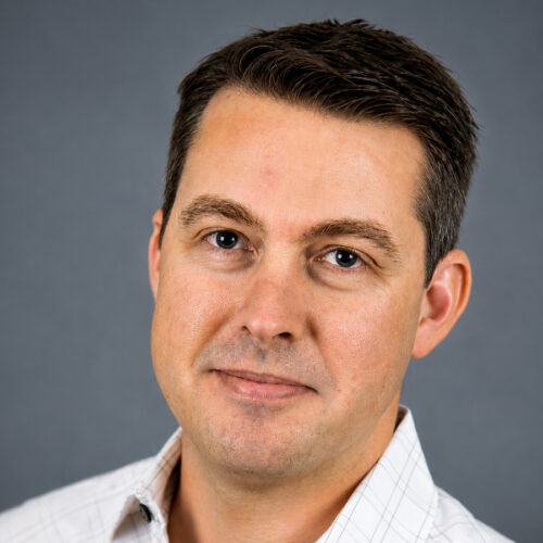 Man with short brown hair wearing a light-colored collared shirt, facing the camera and smiling slightly, with a plain gray background.