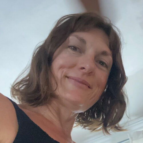 A woman with wavy brown hair smiles at the camera in a close-up selfie, wearing a black top, indoors with soft lighting.