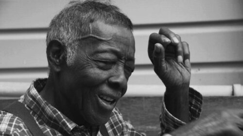 An elderly man with short gray hair and a plaid shirt smiles with his eyes closed, raising one hand near his head.