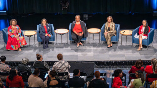 Five women sit on stage in armchairs with small tables between them, facing an audience in a theater or auditorium setting.