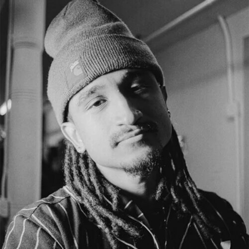 A person with long dreadlocks and a beanie looks at the camera with a neutral expression in a black and white indoor photo.
