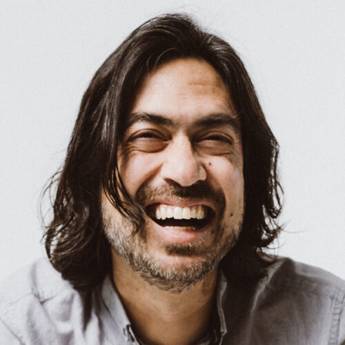 A man with shoulder-length dark hair and a beard is smiling broadly, showing his teeth. He is wearing a light gray collared shirt and appears happy, with a white background behind him.