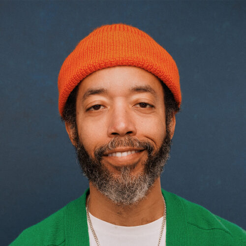 A man with a black and gray beard wearing an orange knit hat, green sweater, and white shirt smiles gently in front of a blue background.