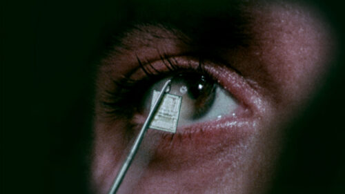 A close-up of a human eye with a small microchip being held near the eyeball by metallic tweezers.