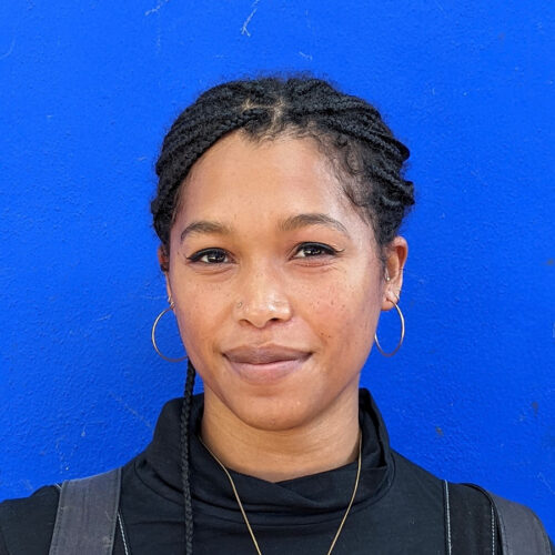 Woman with braided hair, gold hoop earrings, and a black top stands in front of a bright blue wall, facing the camera with a slight smile.