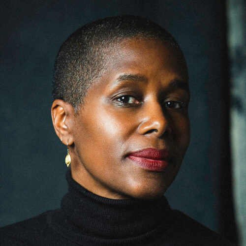 Person with short hair, black turtleneck, and gold earring looks confidently at the camera against a dark background.