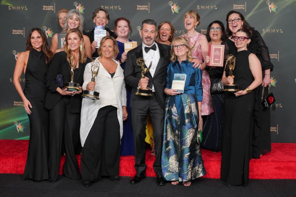 People in formalwear stand on a red carpet holding Emmy awards and books, smiling in front of an official Emmy Awards logo backdrop.