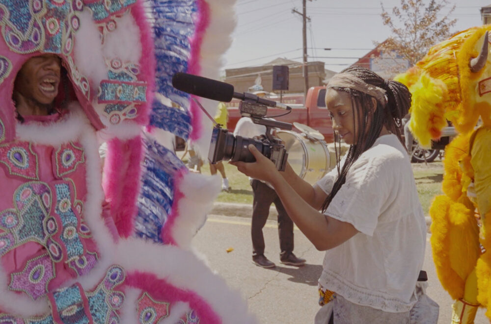 A filmmaker with a handheld camera records a person in an elaborate pink and blue Mardi Gras Indian suit, standing near a person in an orange feathered costume.